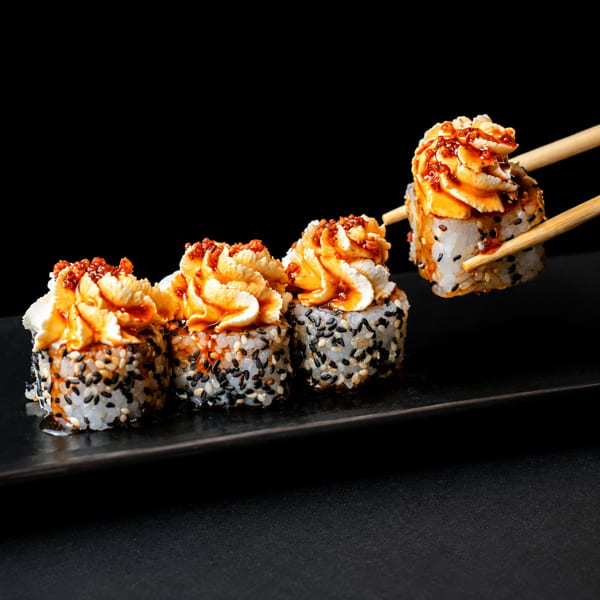 Chopsticks lifting a sesame seed sushi roll topped with creamy sauce and red roe from a black plate.