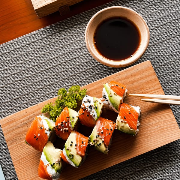 Salmon and avocado sushi rolls with cream cheese, sesame seeds, and soy sauce on a wooden board.
